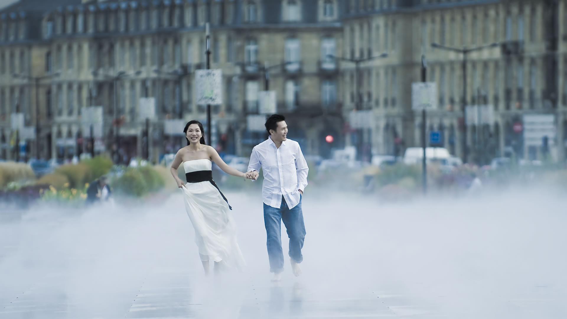 Couple courant main dans la main dans la brume légère du Miroir d'eau de Bordeaux, avec de grands bâtiments en pierre derrière eux
