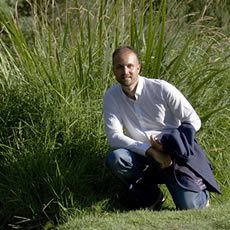 Homme souriant en chemise blanche, accroupi dans un jardin herbeux, une veste bleu marine à la main