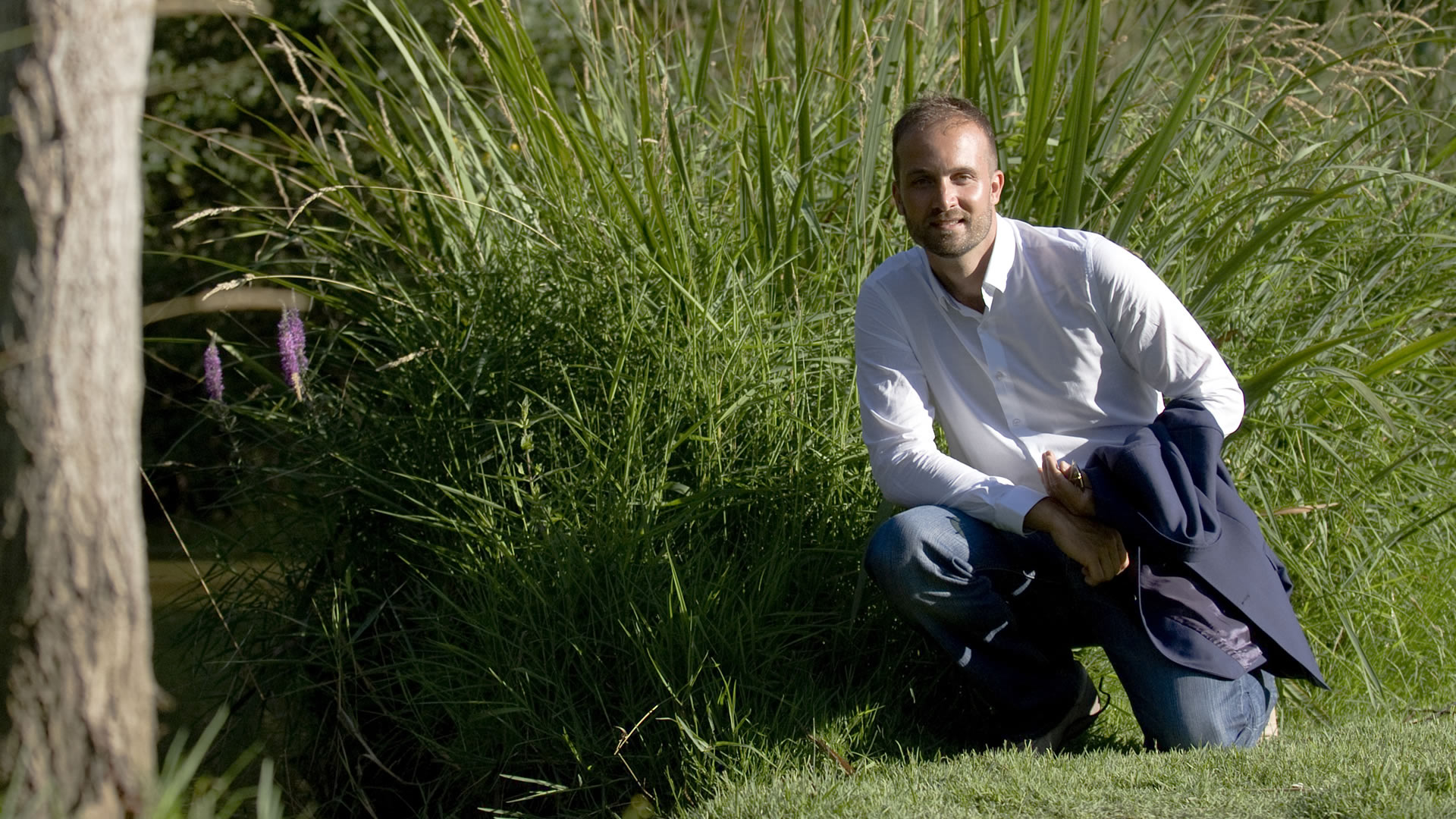 Homme souriant en chemise blanche, accroupi dans un jardin herbeux, une veste bleu marine à la main