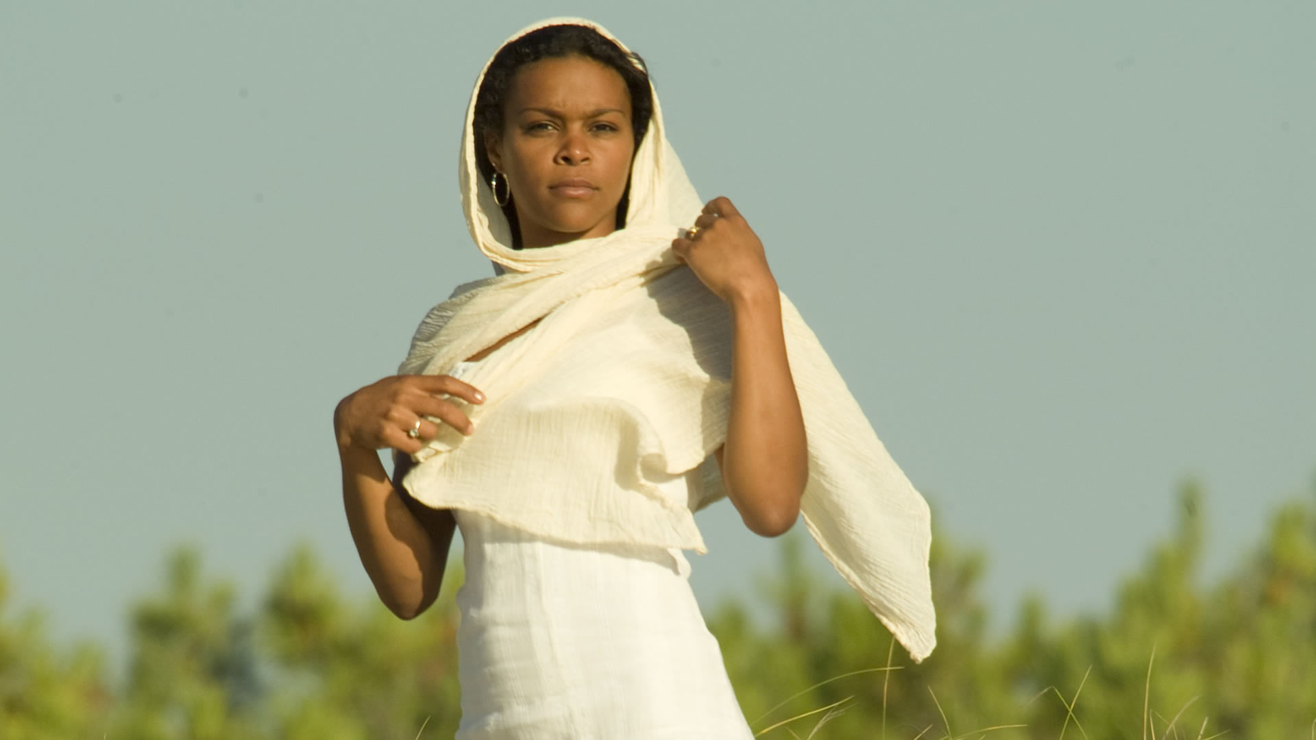 Femme en robe blanche, un foulard crème drapé sur la tête, debout face à un ciel bleu pâle