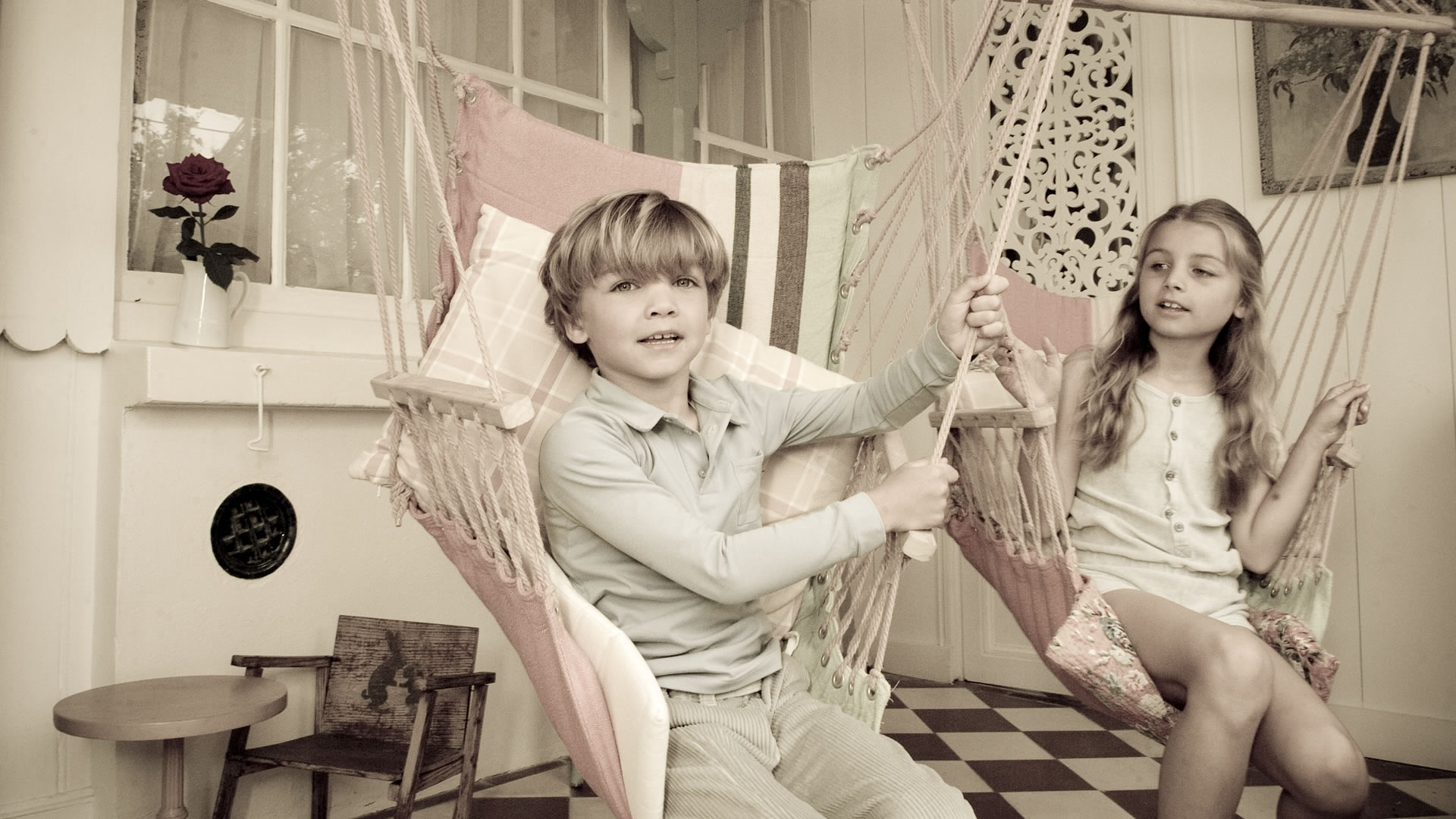 Un garçon et une fille assis dans des fauteuils suspendus rose et crème, dans une pièce lumineuse au style vintage