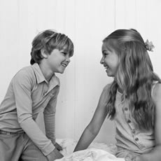Photo en noir et blanc d'un garçon et d'une fille riant, face à face sur un lit devant un mur lambrissé blanc