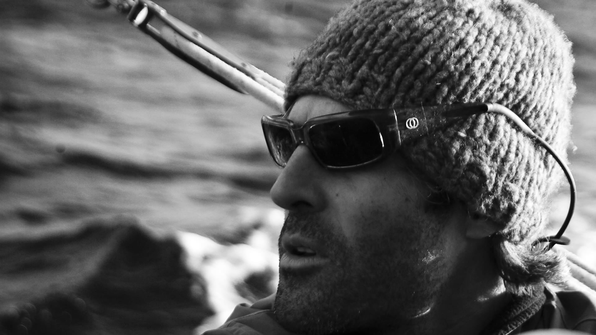Gros plan en noir et blanc d'un homme barbu en bonnet en maille et lunettes de soleil, avec une eau agitée derrière lui